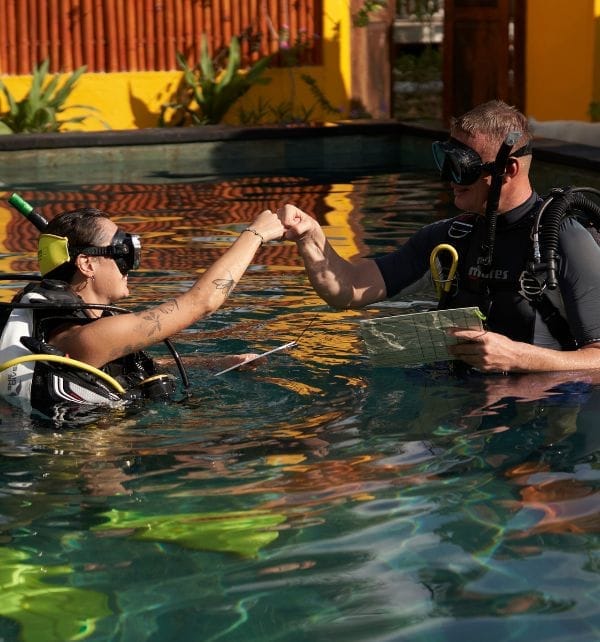 Scuba instructor and student give a fist bump during a training session in a pool at Nusa Lembongan, both in full dive gear.
