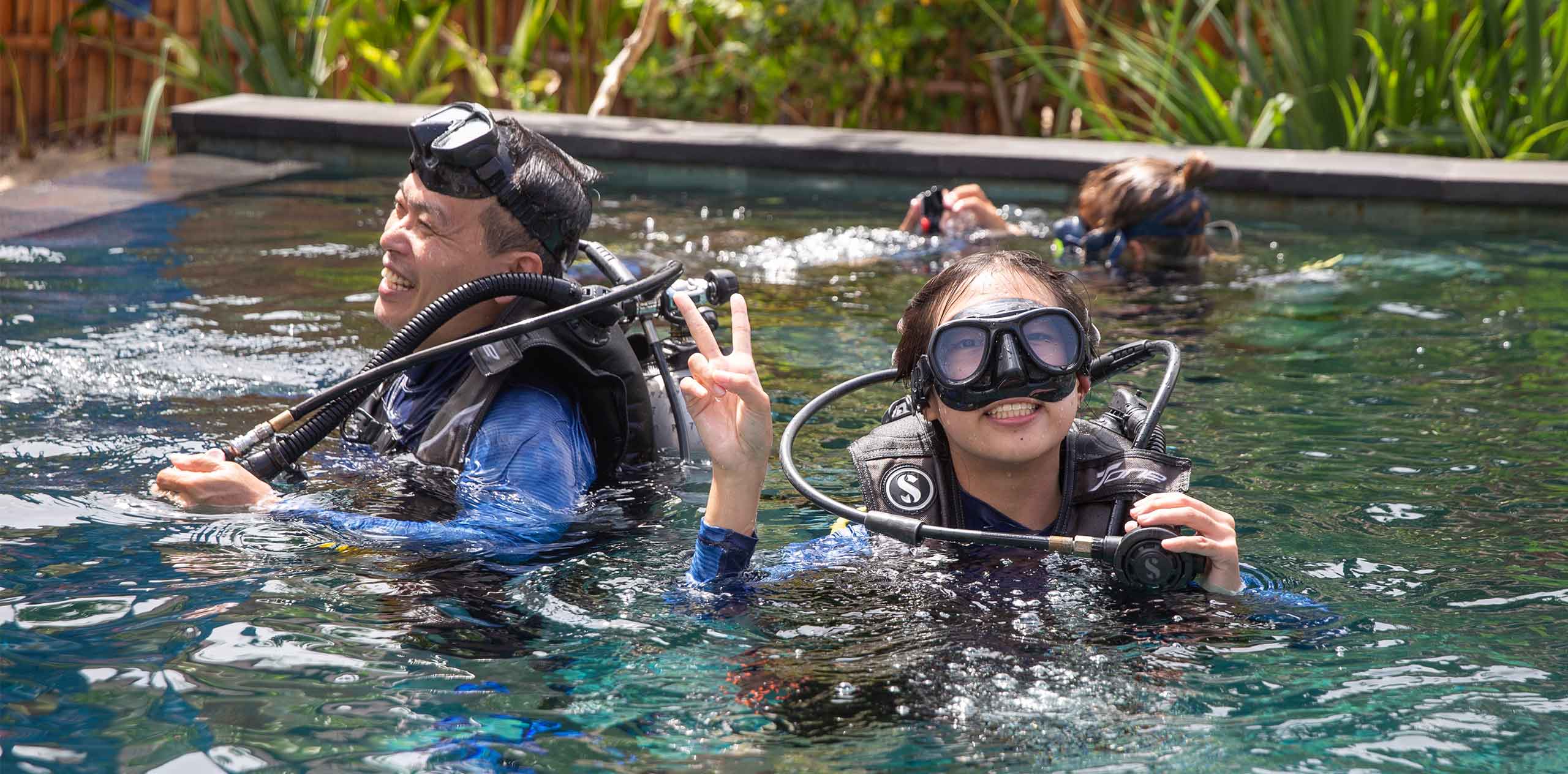 Two people practicing scuba in a training pool on Nusa Lembongan, woman flashing a peace sign while wearing mask and regulator.