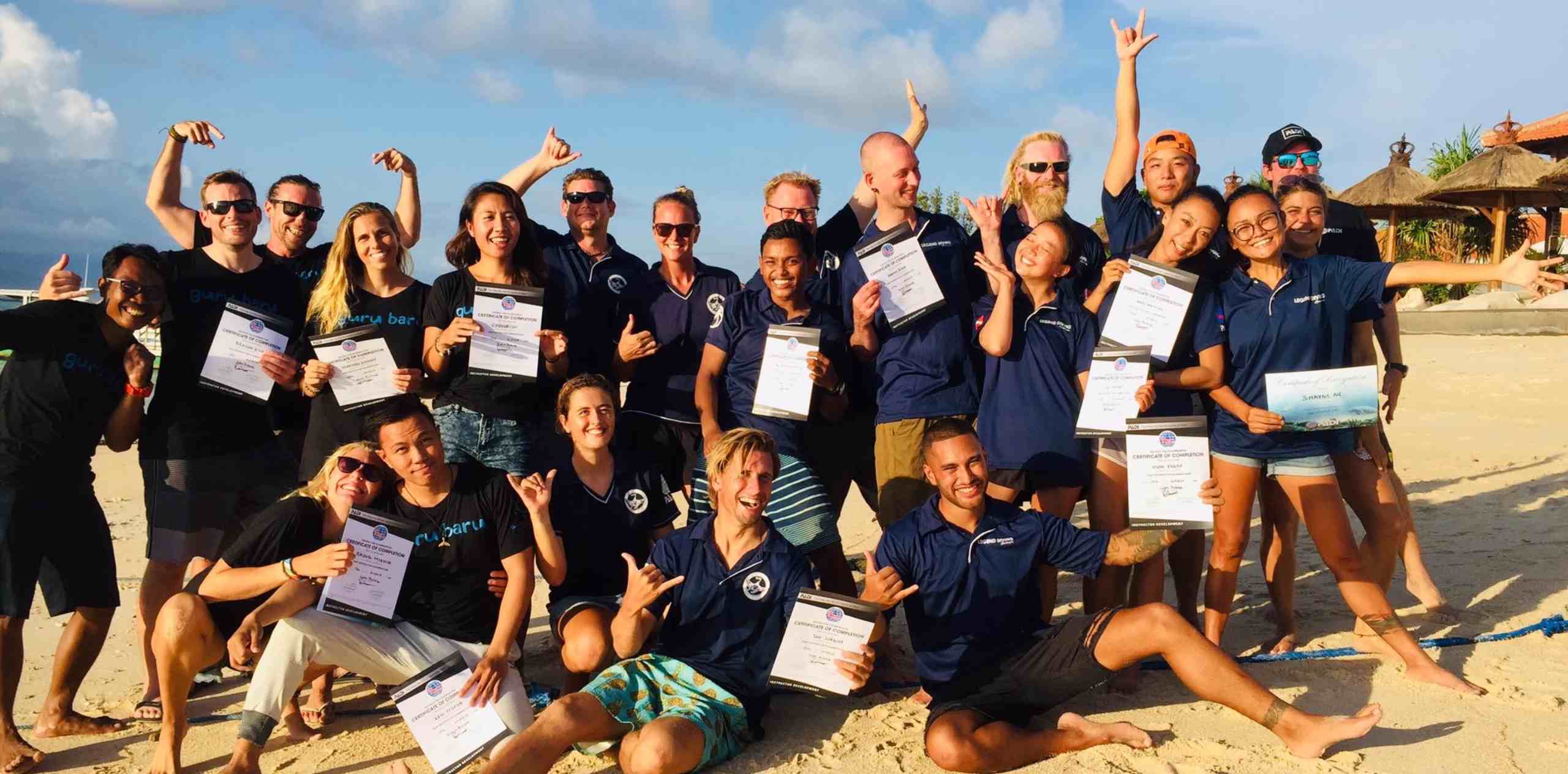 Group of smiling people on Nusa Lembongan beach holding certificates and making celebratory hand signs after a course.