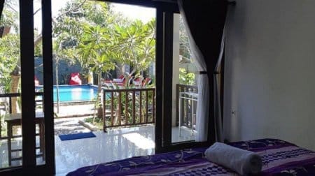 Bedroom with purple bed and rolled towel opening to a balcony framed by tropical plants and a swimming pool.
