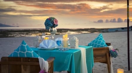 Romantic sunset beach dinner setup on Nusa Lembongan with a turquoise table for two, candles, flowers and ocean backdrop.