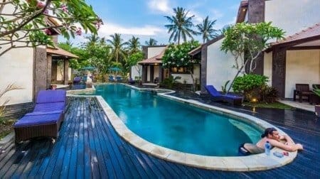 Guest relaxing at a resort pool on Nusa Lembongan, lying at the pool edge with sun loungers, villas and palm trees.