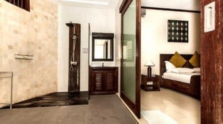 Open-plan Nusa Lembongan hotel room with a walk-in shower on the left and a neatly made bed with pillows visible through a glass partition