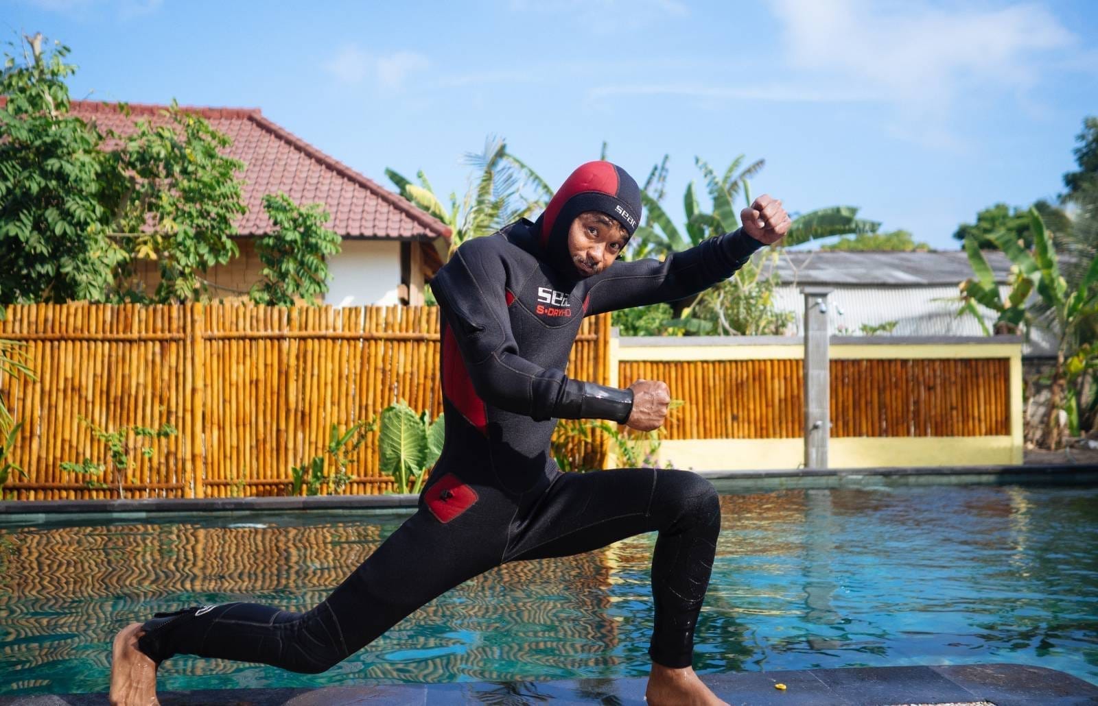 Man in black-and-red wetsuit lunging with fists raised beside a pool on Nusa Lembongan