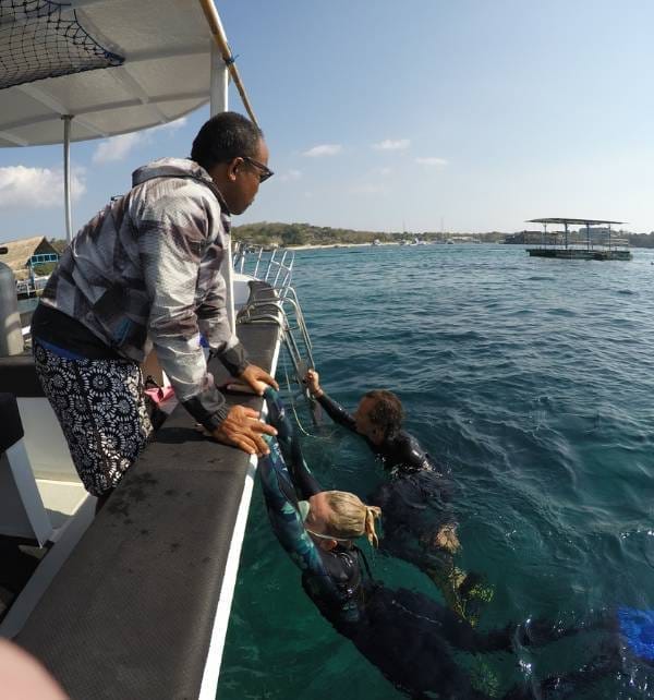 Decompress 2 Two snorkelers in wetsuits climb from the water onto a dive boat as a crew member helps, off Nusa Lembongan.
