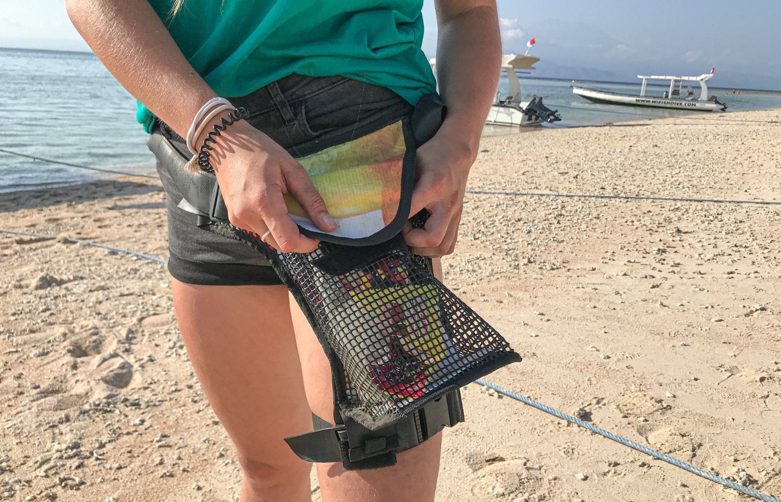 Person on Nusa Lembongan beach fastening a mesh thigh pouch to their leg and placing gear into it, boats in background.