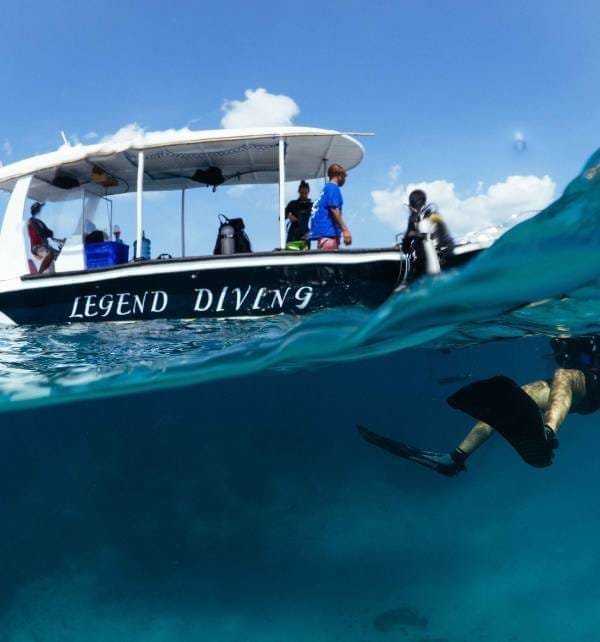 The experience of lifetime 3 Diver with fins swims underwater beside a boat labeled Legend Diving as crew prepare gear off Nusa Lembongan.