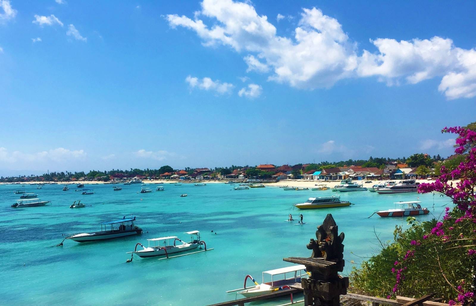 Boats anchored on turquoise water off Nusa Lembongan, paddleboarders near the shore and a coastal village beyond.