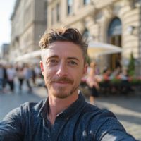 Man with short curly hair, mustache and goatee smiling in a sunlit city street selfie with outdoor cafes blurred behind him.