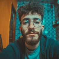 Young man with curly hair, round glasses and beard looking at the camera in a teal-orange lit room with a patterned wall.
