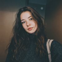 Young woman with long wavy brown hair wearing a dark sweater and a shoulder-strap bag, neutral expression indoors