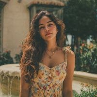 Woman with long wavy brown hair wearing a floral sundress and necklace, standing by a stone fountain and sunlit garden.