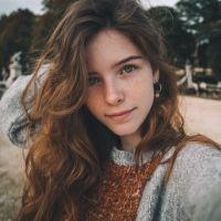 Young woman with long wavy brown hair and freckles wearing sweaters and a hoop earring, park fountain behind her.