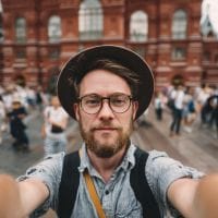 Bearded man with glasses and a hat taking a selfie in a crowded square with a red brick historic building behind him.