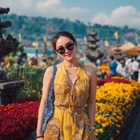 Young woman in a yellow patterned sundress and sunglasses smiling amid bright marigolds with a lakeside temple behind her