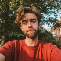 Young man with tousled brown hair and short beard in a red shirt taking a selfie outdoors with trees and houses behind him.