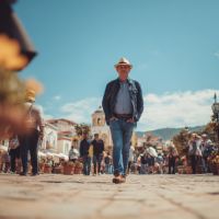 Man wearing a straw hat, sunglasses and denim jacket walking toward the camera on a sunlit cobblestone street lined with crowds and colonial-style buildings.