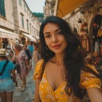 Young woman with long dark hair in a yellow floral dress taking a selfie on a cobblestone street with market stalls.