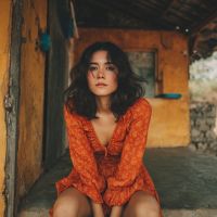 Woman with shoulder-length dark wavy hair wearing an orange patterned dress sits on a stone step in a rustic yellow-walled veranda, gazing at the camera.