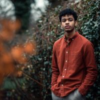 Young man in a rust-colored button-down shirt standing with hands in pockets in front of blurred autumnal foliage, looking at the camera.