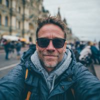 Man with short gray beard and sunglasses smiling at camera, wearing a hoodie and jacket on a busy city street