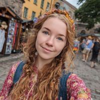 Young woman with long curly red hair, braided strands and floral headband smiles on a cobblestone street with market stalls.