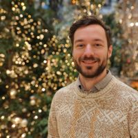 Smiling bearded man in a patterned knit sweater standing before twinkling holiday lights and decorated fir trees.