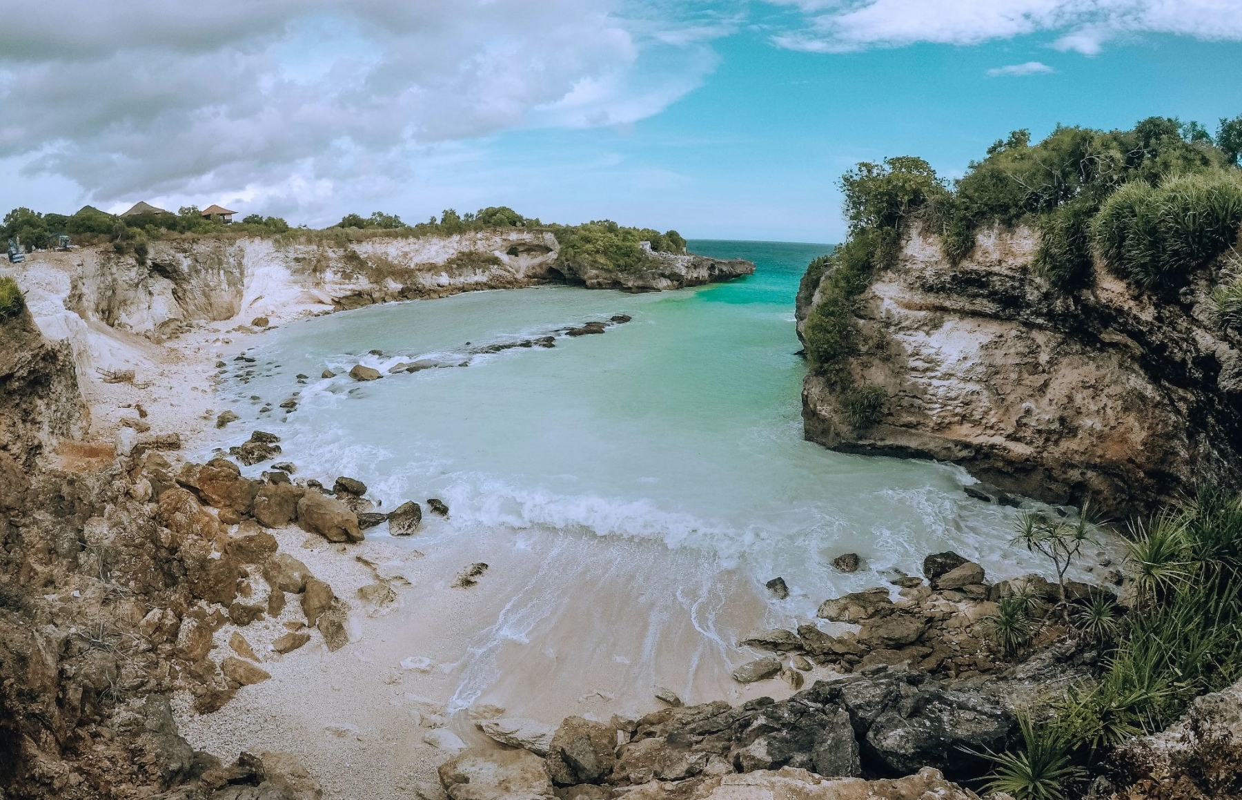 Secluded rocky cove with turquoise water, sandy shore and limestone cliffs topped with green vegetation under a partly cloudy sky
