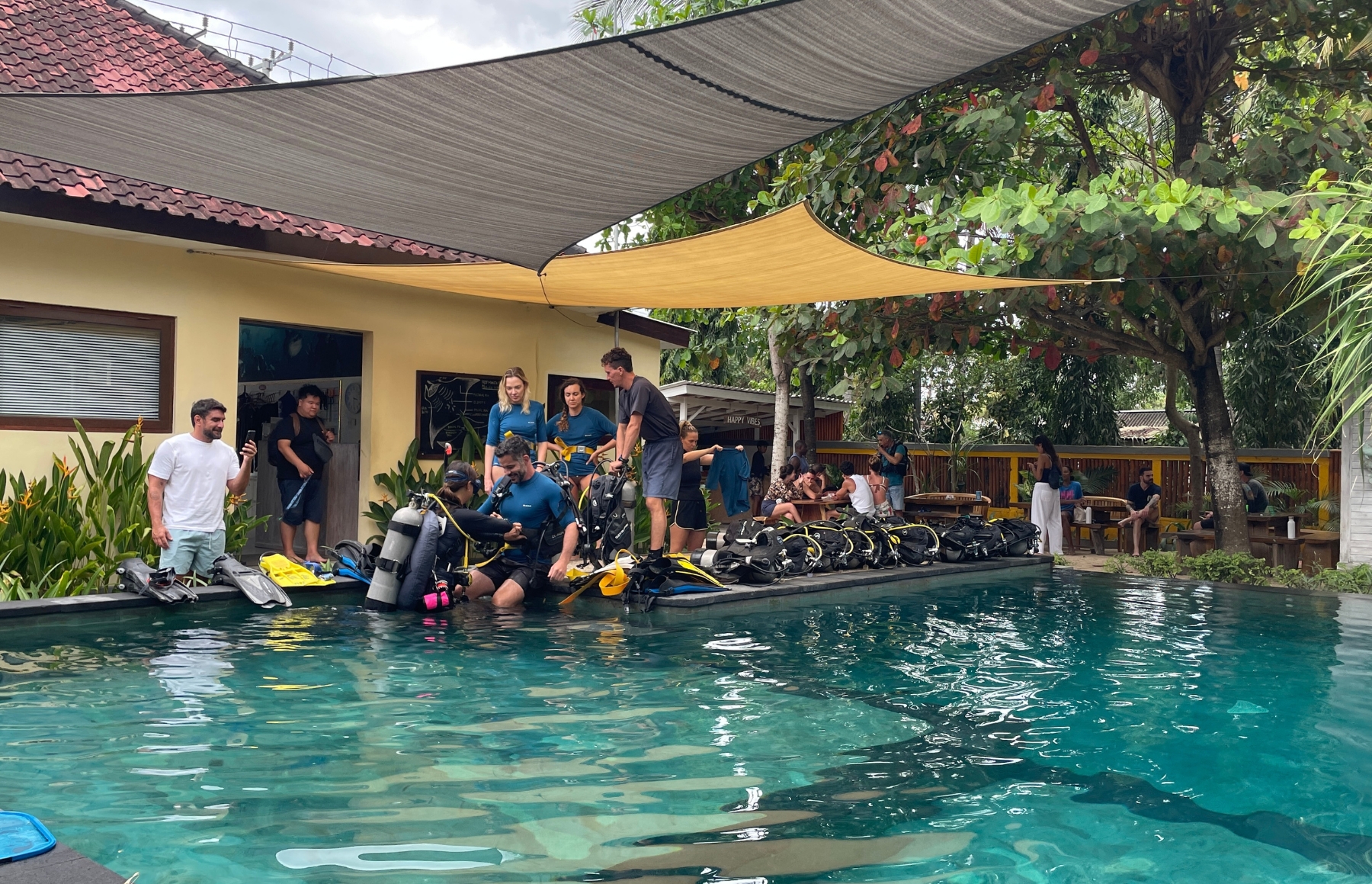Group of divers with scuba gear preparing at a poolside station under shade sails, instructors assisting and others seated nearby.